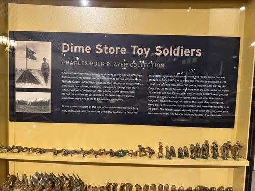 Close-up of the title panel for the toy soldier collection: dark header reading 'Dime Store Toy Soldiers / Charles Polk Player Collection' with two black-and-white archival photos at left (one of a soldier beside a tent and a flag, one of formation) and two columns of explanatory text. Rows of painted soldiers sit on the shelves below.