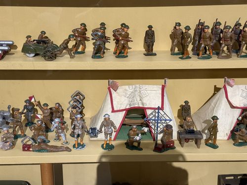 Two shelves of the toy soldier case in close-up. Top shelf has marching infantry, a soldier firing a machine gun from behind a wheeled gun shield, and an officer figure. Lower shelf shows a field-camp scene with two white canvas tents marked 'U.S. SIGNAL CORPS Co A', soldiers operating radio/telegraph gear, a flag-bearer, a kneeling gunner, and a prone soldier.