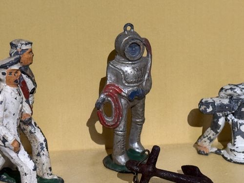 Extreme close-up of three worn toy figures against a tan backdrop: at left a sailor in white uniform with paint chipped off, in the center a silver-painted hard-hat deep-sea diver holding a coiled air hose, and at right another chipped white-uniformed figure crouching. A small rusty anchor prop sits in the foreground.