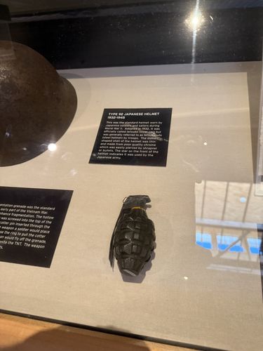Inside a glass artifact case: a rounded steel Japanese WWII helmet at upper left and a green pineapple-pattern fragmentation hand grenade resting on a fabric mat. Two white-on-black label cards are mounted in the case - one titled 'Type 92 Japanese Helmet 1932-1945' and a partly visible one (left) describing a Vietnam-era fragmentation grenade.