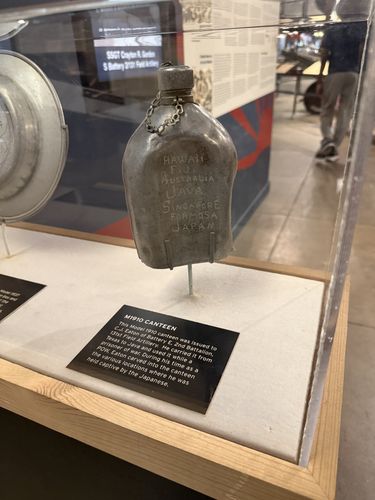 A dented aluminum WWII canteen mounted upright on a stand inside a glass case. Place names are scratched into the metal by hand: HAWAII, FIJI, AUSTRALIA, JAVA, SINGAPORE, FORMOSA, JAPAN. A black label card 'M1910 Canteen' sits below. In the reflected/background gallery a panel reads 'SSGT Crayton R. Gordon / B Battery 2/131 Field Artillery'.
