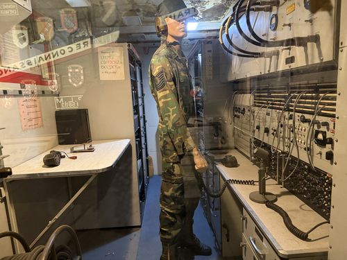A walk-in command/communications shelter diorama seen through glass. A mannequin in 1980s-90s woodland-camouflage BDUs and a helmet stands holding a radio handset; the right wall is a bank of military radio transceivers and patch panels with coiled cords and a desk microphone. The left wall has a folding map table, a field telephone, and posted French-language signs and unit patches/crests.
