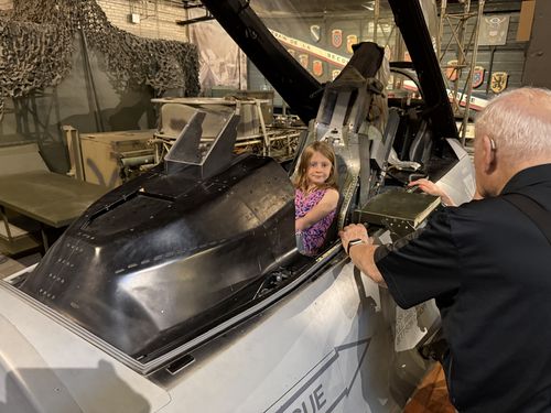 Hands-on exhibit: a girl in a pink top sits inside the open black cockpit of a fighter jet (an F-16-style trainer/mockup) painted in gray and white. An older gray-haired man in a dark polo leans in from the right, lifting the canopy or a panel and apparently explaining the controls. Camouflage netting and other vehicles fill the hangar background; French-language signage is visible behind.
