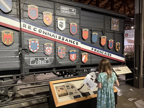 The large dark-painted French WWI/WWII railway boxcar (the 'Forty and Eight' / Merci Train car). A red-white-blue diagonal banner reads 'RECONNAISSANCE FRANCAISE'; mounted around it are painted heraldic shields for French provinces (Maine, Lorraine, Corse, Berry, Auvergne, Touraine, Vendee, Bourgogne, Guienne, etc.), a U.S. eagle crest, and a stenciled capacity sign 'HOMMES 40 / CHEVAUX (EN LONG) 8' plus the railway marking 'P.L.M. J 58113'. A girl in a green dress stands at a glass-topped exhibit table reading a printed sheet in front of the car.