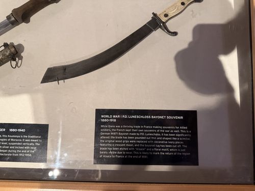 A wall-mounted glass case of blades. The main object is a long curved scimitar-like souvenir blade with a light bone/ivory-handled hilt, displayed point-down, with its shadow falling across the back mat. A black label card (text rotated sideways) titled 'World War I P.D. Luneschloss Bayonet Souvenir 1880-1918' sits beside it; another partial card at upper left describes a Moroccan dagger (1880-1940).