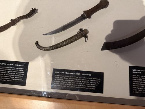 Another view into the same blade case. A curved Moroccan koummya dagger with an ornate engraved metal scabbard lies beside its straight pointed knife blade with a wooden grip. Above is a carved wooden-handled blade, and below the edge of the scimitar souvenir is visible. Two sideways black label cards are readable: 'Moroccan Koummya Dagger 1880-1940' and the edge of a 'Letter Opener 1918-1920' card.