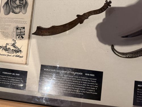 Close view of a rusty, curved 'trench art' letter opener shaped like a miniature sword, displayed on the case's cream mat with a strong shadow. Above it is a yellowed open magazine page (a 'Gro-Pup' dog food ad with a dog illustration) dated February 28, 1949. The black label card (sideways) is titled 'World War I Souvenir Letter Opener 1918-1920'.