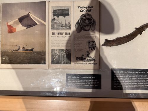 View of the same case showing the open vintage LIFE magazine (February 28, 1949) propped inside: a top photo of a ship at sea flying a large French tricolor flag, a middle spread headlined 'THE "MERCI" TRAIN / France sends 49 boxes full of gifts to the U.S.', and the lower 'Gro-Pup' dog food ad ('Can't help lovin' GRO-PUP'). The curved copper trench-art letter opener and the edge of its label card are at lower right.