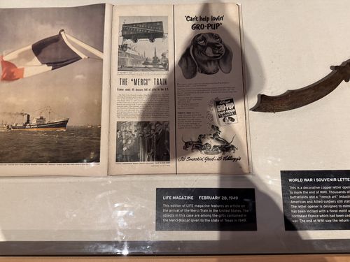 A framed display case about the post-WWII 'Merci Train' (French Gratitude Train of 1949). It holds an open copy of LIFE magazine showing a photo of a ship flying the French tricolor and an article titled 'THE "MERCI" TRAIN,' plus a vintage 'Can't help lovin' GRO-PUP' Kellogg dog-food advertisement on the facing page. Below is a curved WWI souvenir copper letter opener. Two small typed placards label the items.