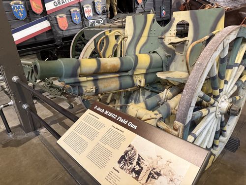 The actual 3-inch M1902 Field Gun, painted in a yellow-green-and-blue WWI 'dazzle' style camouflage including the spoked wheels, with its open breech and recoil cylinders visible. Behind it stands the wooden 'Merci Train' boxcar covered in colorful French regional coats of arms, with a French tricolor and 'GRATI[TUDE]' lettering. The matching placard sits on a stand in front.