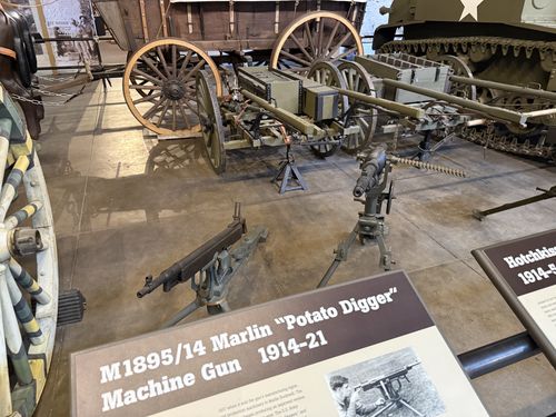 The actual Marlin M1895/14 'Potato Digger' machine gun mounted low on its tripod in the foreground, with a Hotchkiss machine gun on a taller tripod beside it (partial placard reading 'Hotchkiss... 1914-5...'). Behind them sit horse-drawn artillery wagons and limbers, and the tracks of an M-series light tank are visible at right.