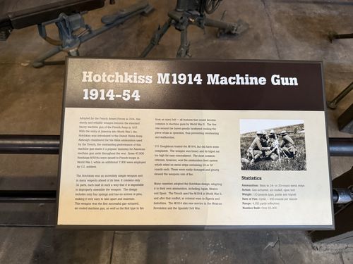 A clear, well-lit placard for the Hotchkiss M1914 Machine Gun, with a black-and-white photo of a French gun crew and a Statistics box. The actual gun's tripod legs are visible above the sign.