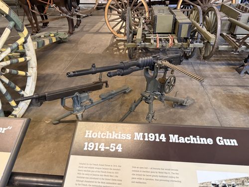 The actual Hotchkiss M1914 machine gun on its tripod, with its distinctive ribbed barrel and a rigid metal ammunition strip hanging from the feed. The Marlin 'Potato Digger' is to the left, and horses, wagons, and artillery fill the background. The Hotchkiss placard is in the foreground.