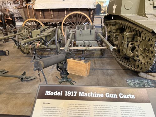 The actual Model 1917 machine gun carts: a wooden gun cart on the left and a green ammunition cart in the center, with a machine gun mounted on a tripod and a wooden ammo chest set in front. A horse harness and a covered Conestoga-style wagon are behind, and the treads of the M3A1 Stuart tank fill the right side. The matching placard is in the foreground.