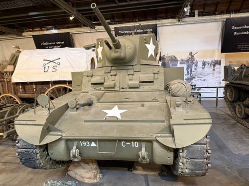 A strong head-on photo of the actual M3A1 Stuart light tank, olive drab with white stars on the turret and hull and front markings '193' (with a triangle) and 'C-10'. Its 37mm gun points up. Behind it: a covered wagon flying a white cavalry guidon with crossed rifles and 'U.S.', and large wall banners -- one reading 'I shall never retreat or surrender!' and another 'The troops of other states have their reputations to make,' plus a black banner about remembering and a large photo of an amphibious/D-Day-style landing. A tan half-track is at far right.