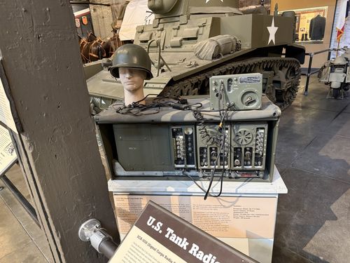 The actual U.S. tank radio display: an olive-drab SCR-508 radio set (transmitter and receiver modules) on its tray, with a separate remote/control box on top, coiled cables, and a mannequin head wearing an M1 helmet to represent the crewman. Behind it is the M3A1 Stuart tank, and a tan Afrika-Korps-style half-track is at far right. The U.S. Tank Radio Equipment placard is in the foreground.