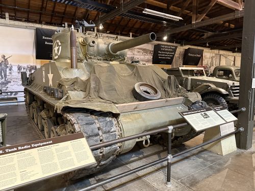 Three-quarter front view of an M4A3(105) Sherman tank in the Great Hall, with a long gun barrel, white star, spare track links draped over the hull, and camouflage netting. Background banners read 'Victory or Death' and quote Robert E. Lee.