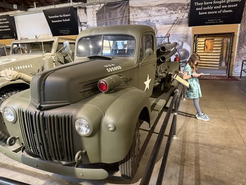 Olive-drab WWII Ford truck marked 'USA 505650' with twin headlights and a red warning light, towing an artillery-style apparatus. A girl (Maxine) in a green dress stands at right reading a clipboard/phone next to the display rail and a doorway with log-cabin backdrop.