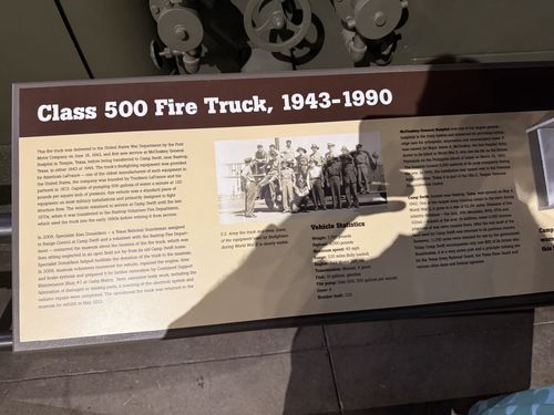 Placard titled 'Class 500 Fire Truck, 1943-1990' with restoration history, a historic photo of an Army fire truck and crew, a Vehicle Statistics box, and a partly-glare-obscured column on McCloskey General Hospital and Camp Swift.