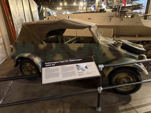 The captured Volkswagen Type 82 Kubelwagen itself: a camouflaged convertible with canvas top up and a spare tire on the back, its placard mounted in front. Behind it sits the desert-tan SdKfz 251 halftrack, with aircraft and flags overhead in the Great Hall.