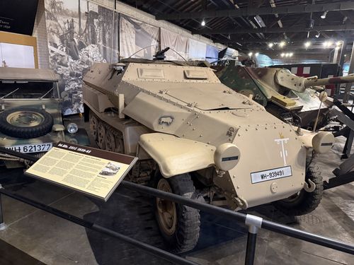 Desert-tan German SdKfz 251C halftrack marked 'WH-939491' with an Afrika Korps palm-tree-and-swastika symbol on the front, an open-topped armored body and a forward MG mount. A camouflaged Kubelwagen (WH 621377) sits to the left and the Hetzer tank destroyer is behind to the right.