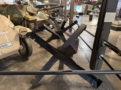 A black steel 'hedgehog' anti-tank obstacle (three crossed I-beams) displayed on the floor of the Great Hall. To the left are the tan SdKfz 251 halftrack (...939491) and the camouflaged Hetzer tank destroyer; a small placard is mounted on the post at right.