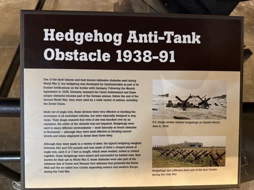 Placard titled 'Hedgehog Anti-Tank Obstacle 1938-91' with body text and two photos: U.S. troops sheltering behind hedgehogs on Omaha Beach June 6, 1944, and hedgehogs/pillboxes forming part of the Iron Curtain during the Cold War.