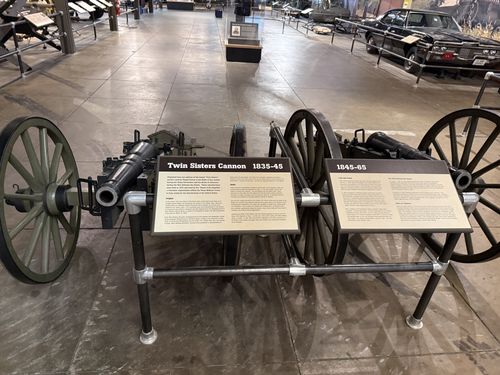Wider shot of the same Twin Sisters display: two green-wheeled replica cannons mounted side by side on a black tubular steel frame, with the 'Twin Sisters Cannon 1835-45' placard and the adjacent '1845-65' placard between them. The Great Hall stretches behind with other vehicles including a black sedan/limousine on the right.
