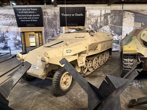 A sand-colored German SdKfz 251 half-track in the Great Hall, marked with vehicle number 833431 on the front and a German cross on the side, displayed in front of large banners reading 'I rely upon our Texans...' (Robert E. Lee) and 'Victory or Death.' (William Barrett Travis). The dark Jagdpanzer 38 Hetzer is just visible at right.