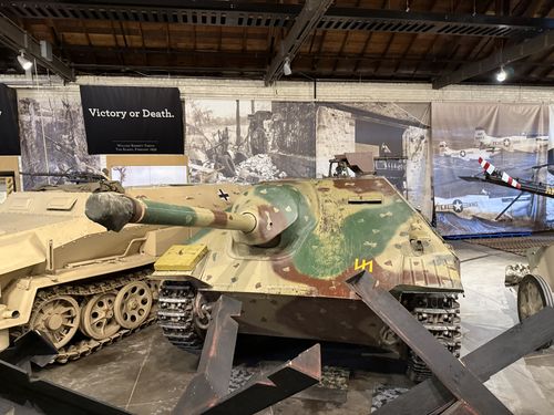 The Jagdpanzer 38 Hetzer itself in the Great Hall, painted in green/brown/tan camouflage with a long gun barrel, a German cross and yellow stowage box, displayed behind anti-tank steel hedgehog obstacles. The tan SdKfz 251 half-track is at left and aircraft/banners are in the background.