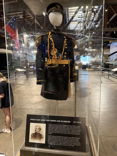 The display case holding General Mabry's dress uniform: a dark blue/black frock coat with brass buttons, gold shoulder cords and aiguillette, a sash, a peaked cap on a headform, and a sword with scabbard. A Confederate-style flag is at left and the museum's tanks and aircraft are reflected/visible in the background. The placard from index 49 is at the base of the case.