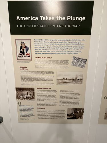 Large wall panel titled 'America Takes the Plunge - The United States Enters the War', covering 1915-1917 events, with an 'I Want You for U.S. Army' Uncle Sam poster, a German newspaper image, a U-boat photo, and sections headed 'He Kept Us Out of War', 'Telegrams and U-Boats', 'America Declares War', and 'Mobilization'.