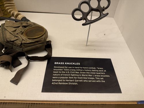 Inside a display case: a black metal set of brass knuckles mounted on a stand, next to a canvas/leather gas-mask bag with a round metal canister, and a small black 'Brass Knuckles' label.