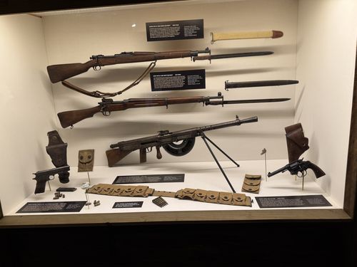 Wide overview shot of a glass display case of WWI-era US small arms: two bolt-action rifles with bayonets and scabbards at top, a light machine gun (Chauchat automatic rifle) on a bipod in the middle, two pistols (a semi-auto and a revolver) in holsters at the sides, ammunition belts, magazines and clips below, each with its own small black placard.