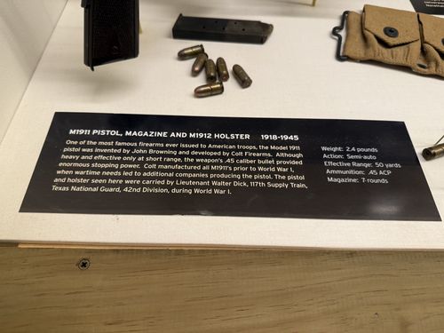 Close-up of the placard for the M1911 Pistol, with the pistol's magazine and loose .45 ACP cartridges visible above the sign.