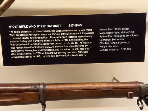 Close-up of the placard for the M1917 Rifle and M1917 Bayonet, with the wood-and-steel rifle barrel resting on its mounts below the sign.