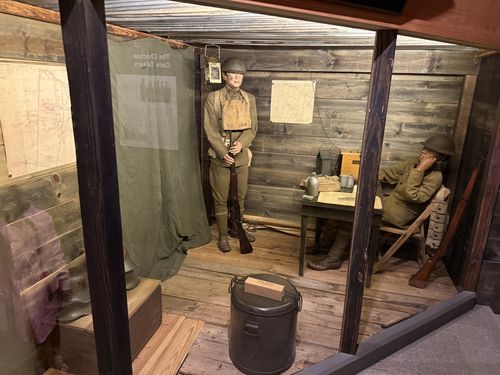 Recreated WWI 'Post of Command' bunker diorama viewed through the display opening. A standing doughboy mannequin in helmet and pack holds a bolt-action rifle; a second seated mannequin slouches at a rough plank table with a candlestick field telephone, mess gear and a rifle leaning beside him. Walls are rough timber with situation maps tacked up. A canvas curtain on the left is back-printed with 'The Choctaw Code Talkers' (text reads mirror-reversed from this angle). A marmite/insulated food can sits in the foreground.