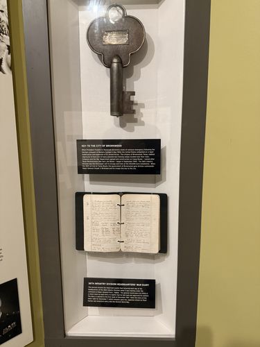 Tall vertical display case with an oversized ornamental iron skeleton key mounted above an open hand-written notebook. Two black placards label the two objects.