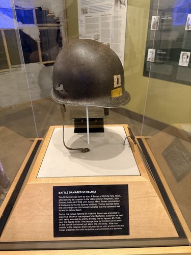 Glass display case holding a battle-damaged olive-drab M1 steel helmet with a captain's bar painted on the front and a ragged shrapnel hole torn through the left side, on a stand above a black label.