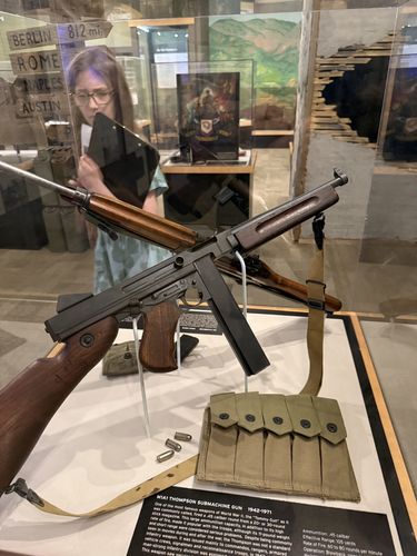 Open display case with an M1A1 Thompson submachine gun (the 'Tommy Gun') mounted at an angle on a stand, a five-cell magazine pouch and loose .45 cartridges below it, and a second rifle behind. Maxine is visible behind the case reading the contents; a distance signpost reading 'BERLIN 812 mi / ROME / NAPLES / AUSTIN' is in the background.