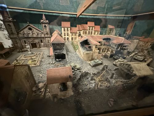 Detailed scale diorama inside a glass case depicting a wrecked Italian town square: a damaged stone church, a fountain, multi-story houses with red tile roofs, a courtyard building, rubble piles, tanks and tiny soldier figures fighting amid the ruins. Painted backdrop behind. Sharp, relatively low glare.