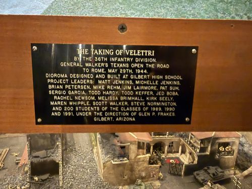 Close-up of a brass plaque mounted above a diorama. Through the bottom of the frame you can see the diorama itself: a battered Italian town with red-tiled roofs, rubble, soldiers, a tank, and a banner bearing a swastika.