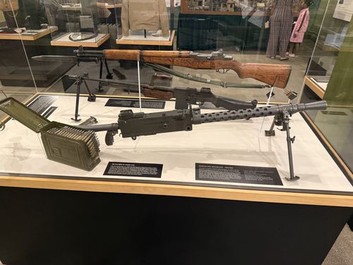 Glass display case of WWII small arms and crew-served weapons: an M1 Garand rifle with bayonet at the back, a light machine gun on a bipod in the middle, a heavy machine gun (likely a .30 caliber Browning) on a tripod with a green ammunition can and linked belt to the left front. Black object labels sit in front of each. Another visitor is visible behind the glass.