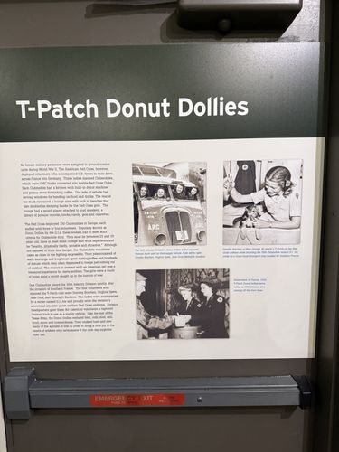 Wall panel titled 'T-Patch Donut Dollies' mounted on what appears to be an emergency-exit door, with three black-and-white photos of Red Cross women, including one holding a kitten by a Clubmobile, and a column of text.