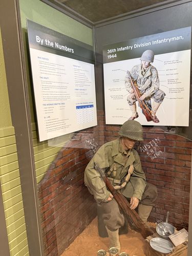 A walk-in display alcove. On the left and back wall are two panels: 'By the Numbers' (statistics) and '36th Infantry Division Infantryman, 1944' with a large illustrated, labeled diagram of a kneeling soldier and his gear. In the foreground, a life-size mannequin of a 1944 infantryman in helmet and field uniform kneels against a brick wall holding an M1 Garand, with mess gear and equipment on the ground. Graffiti on the brick reads 'AUSTIN 5/8'.