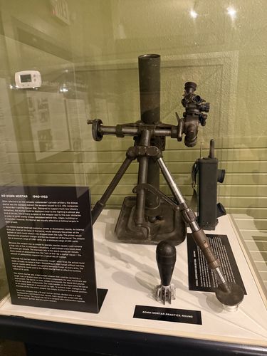 Glass case displaying an M2 60mm mortar set up on its bipod and baseplate, with a sighting unit attached, a field telephone/radio to the right, and a finned practice round and other accessories on the floor of the case. A black object label sits at lower left and a separate small label reads '60MM MORTAR PRACTICE ROUND.'