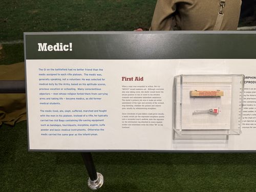 Angled green-and-cream panel titled 'Medic!' with a 'First Aid' section, beside a clear acrylic case holding a glass morphine syrette/ampoule and small box. A mannequin in olive uniform stands behind. Right edge of the panel ('Morphine...') is cut off.