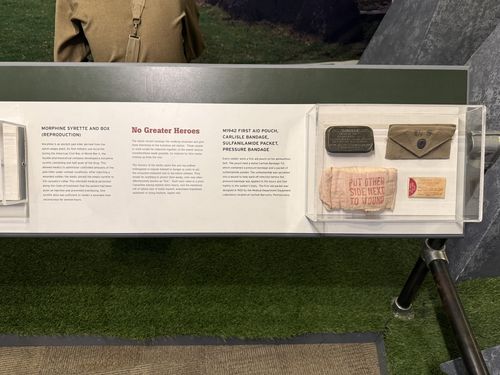 Continuation of the same long panel to the right, headed 'No Greater Heroes', with sub-labels 'Morphine Syrette and Box (Reproduction)' and 'M1942 First Aid Pouch, Carlisle Bandage, Sulfanilamide Packet, Pressure Bandage'. The case holds a green metal tin, a khaki U.S. first-aid pouch, and a folded pressure bandage stamped 'PUT OTHER SIDE NEXT TO WOUND'.