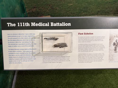 Angled panel titled 'The 111th Medical Battalion' with a 'First Echelon' section. Center acrylic case displays jagged steel shell fragments. A small archival photo of a medic is at the right edge.