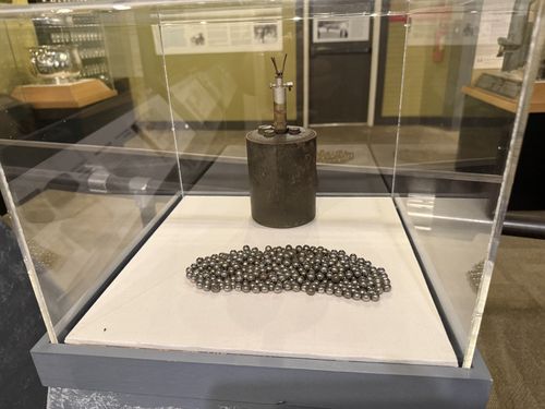 Clear acrylic display case containing the actual German S-mine: a rusted metal cylinder with a three-pronged pressure fuse on top, sitting behind a pile of the small steel shrapnel balls it would scatter. Other exhibits and a glass-bottle display are reflected/visible in the background.
